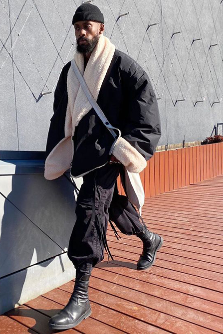 Greg Ntore wearing a beanie hat, long coat, and boots poses against a geometric architectural backdrop 
                            with orange accents.