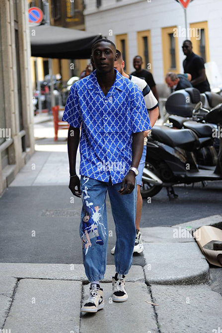 Khaby Lame wearing a blue patterned shirt, distressed jeans, and sneakers walks down a street lined 
                            with motorcycles and buildings in the background.